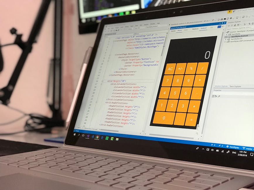 A laptop computer placed on a desk displaying a coding editor alongside a large mobile calculator app with orange buttons for basic arithmetic functions, in an indoor workspace. In the background, a c
