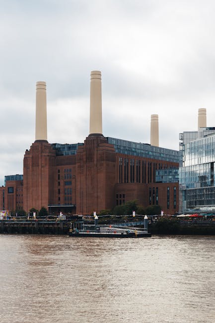 A large brick building with multiple tall, white chimneys rising above it, situated on the bank of a river. The structure appears to be an industrial or historical site, possibly a former power statio