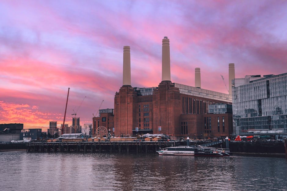 A large brick building with multiple tall, white chimneys rising above it, situated on the bank of a river. The structure appears to be an industrial or historical site, possibly a former power statio